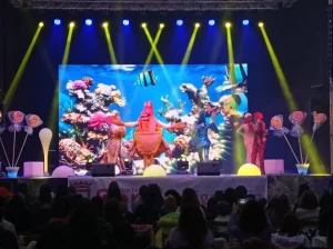 Escenario de un concierto infantil de Gisela Kids, con artistas disfrazados y un pez decorativo en el fondo.