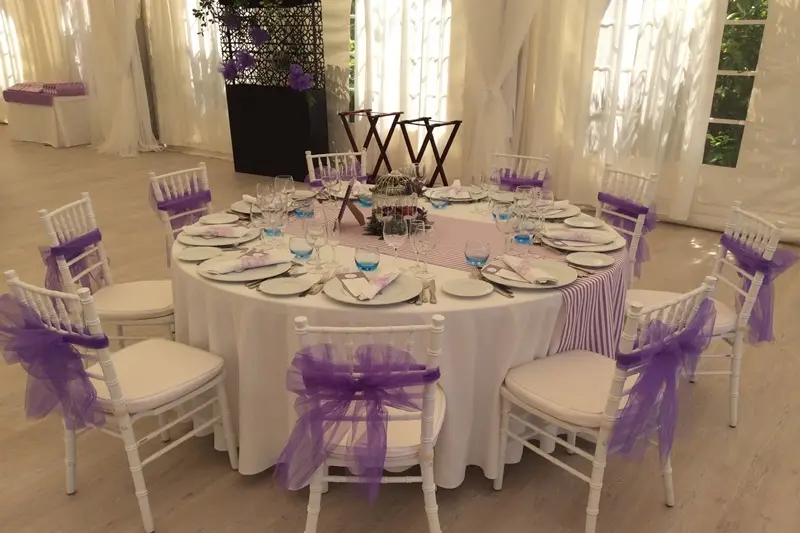 Mesa decorada con sillas moradas y blancas, adornadas con un lazo morado para un evento privado.