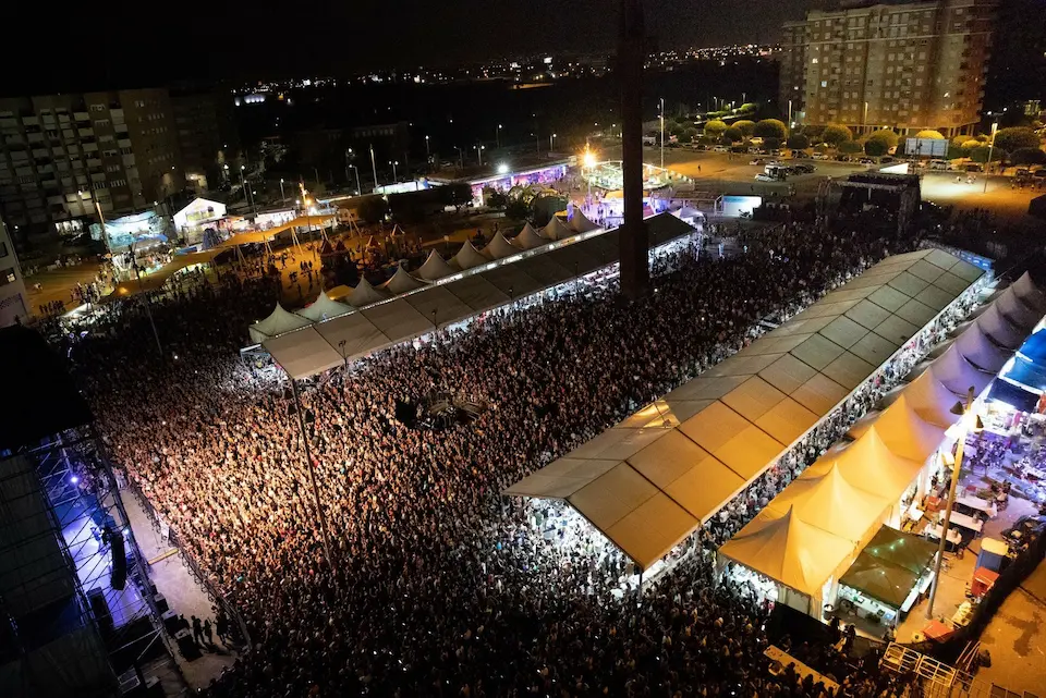 Gran cantidad de asistentes en un concierto, con luces brillantes y un escenario visible al fondo.