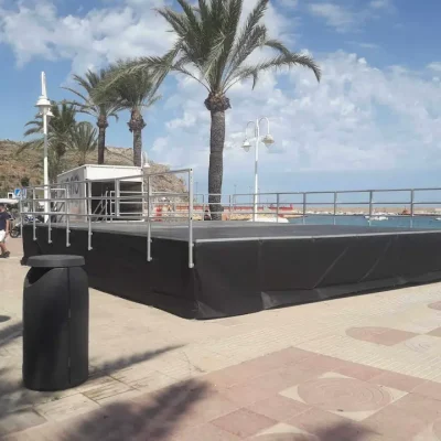 Escenario montado en la playa, rodeado de palmeras, listo para un evento al aire libre.