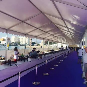 Mesa larga con personas sentadas alrededor, bajo una carpa azul en un ambiente al aire libre.