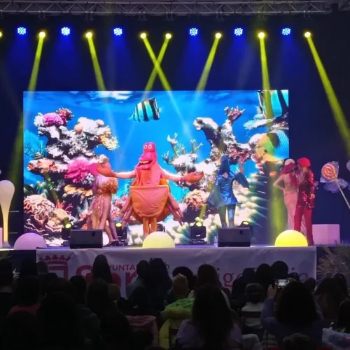 Escenario de un concierto infantil de Gisela Kids, con artistas disfrazados y un pez decorativo en el fondo.