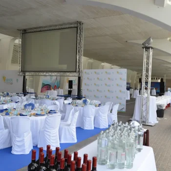 Mesa amplia de color blanco en un congreso, rodeada de sillas y con material informativo sobre la superficie.
