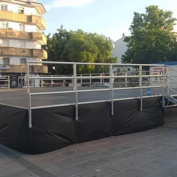 Escenario de aluminio con barandilla metálica y una gran lona negra cubriendo parte del fondo.