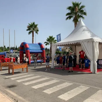 Grupo de personas reunidas alrededor de una carpa con un banner rojo y azul en un evento deportivo.