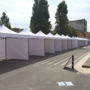 Vista de una feria con carpas blancas en la calle, rodeadas de árboles verdes en el fondo.