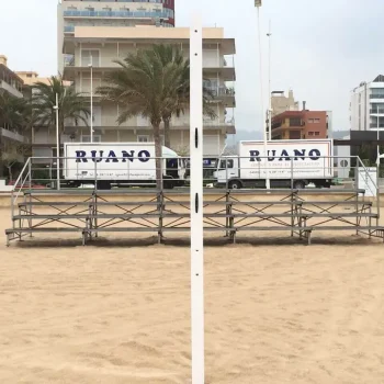 Gradas frente a una cancha de voleibol, con camiones estacionados con el logo de "Ruano Group" en el fondo.