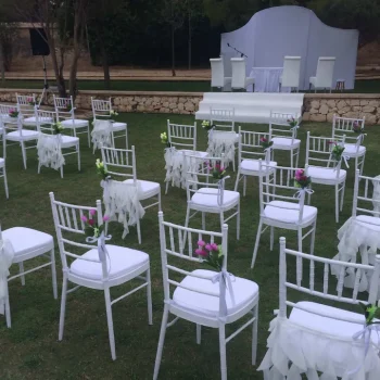 Sillas blancas dispuestas en un jardín para una ceremonia de boda al aire libre, rodeadas de flores y naturaleza.