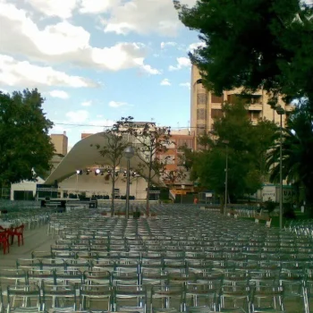 Sillas de metal alineadas frente a un escenario, rodeadas de un gran árbol frondoso.