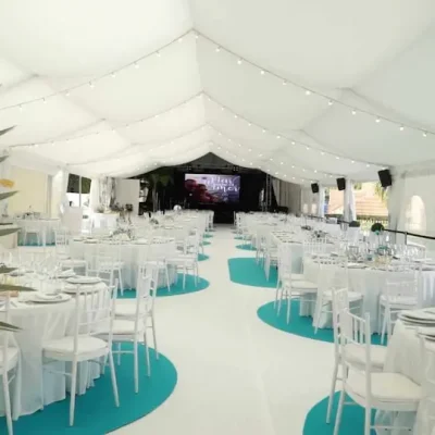 Carpa grande con mesas y sillas blancas y moquetas azules dispuestas para un evento.