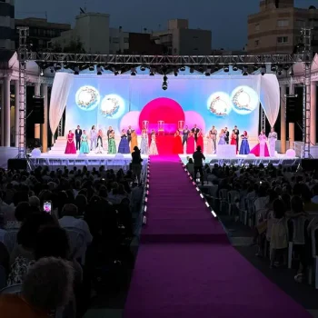 Gran escenario con una multitud observando un certamen de belleza, luces brillantes y decoraciones coloridas.