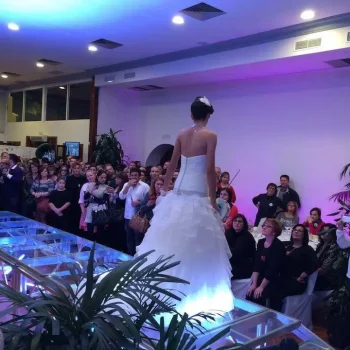 Escenario con rampa, donde una mujer vestida elegantemente se encuentra en una plataforma de vidrio ante un público expectante.
