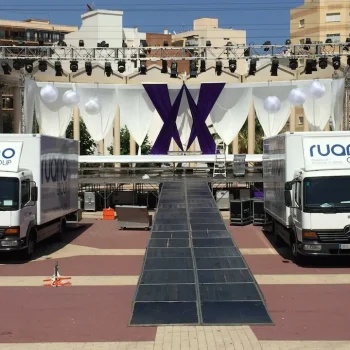 Dos camiones con el logo de “Ruano Group” estacionados frente a un escenario iluminado con luces blancas y una rampa de acceso visible.
