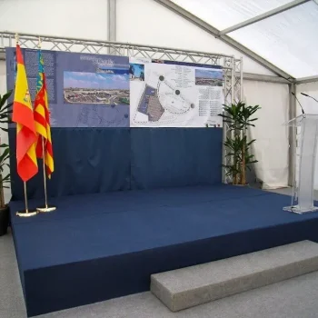 Tarima con un podio y banderas de España dentro de una carpa, lista para un evento o presentación.