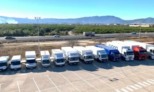 Vista aérea de 15 camiones de una empresa de eventos, estacionados en fila en un aparcamiento amplio.